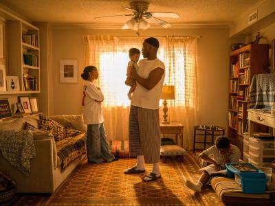 A family scene in a cozy living room with warm lighting. A man holds a child while talking to a woman. Another child is seated on the floor, engaged with a book. The room features a sofa, bookshelves, and a decorative rug.