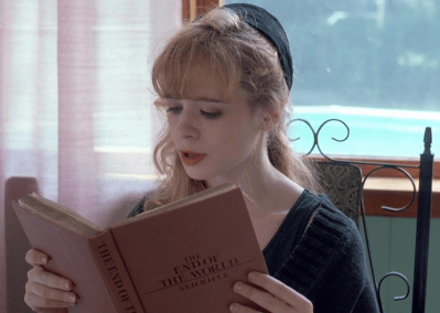 A young woman with long hair and a black headscarf reads a book titled 'The End of the World' while sitting indoors near a window.