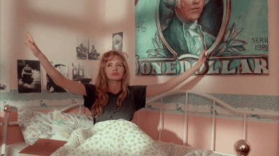 A young woman sitting up in bed with her arms raised, surrounded by pastel-colored walls and a mural depicting a historical figure.