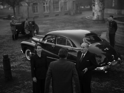 A black and white scene showing two people conversing in front of a vintage car, with a man in a suit and a woman in a dark outfit. In the background, another vintage vehicle and a person in uniform can be seen.