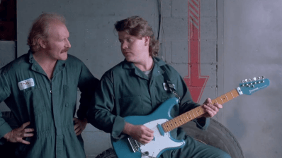 Two male mechanics in green jumpsuits are engaging in conversation, one playing a blue electric guitar while the other looks on.