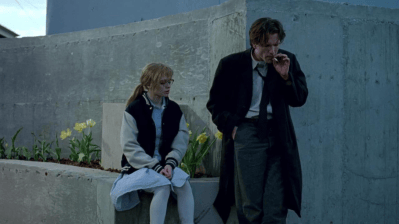 A woman in a varsity jacket sits on a concrete ledge while a man in a black coat stands nearby, smoking. They are surrounded by a minimalist urban setting with yellow flowers in the foreground.