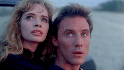 A man and a woman looking up with expressions of concern or awe, against a twilight background, seen from inside a car.