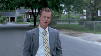 A man in a gray suit and a patterned tie standing on a quiet street with trees in the background.