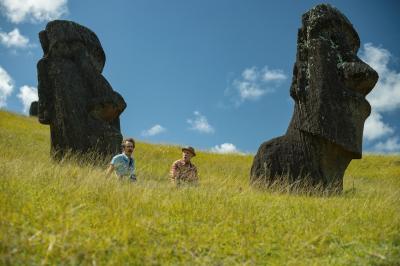 Two people standing in a grassy landscape with two large Moai statues under a blue sky with clouds.
